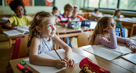 Young girl sitting at a desk in school listening to a teacher while taking notes with a pencil
