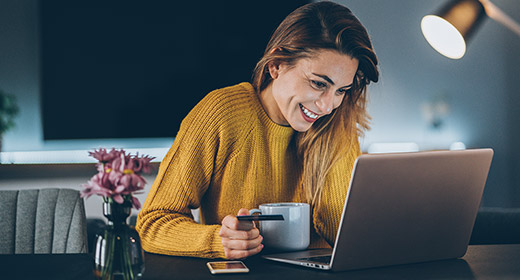 Woman smiling while online shopping at laptop on a desk and hold a coffee mug and credit card