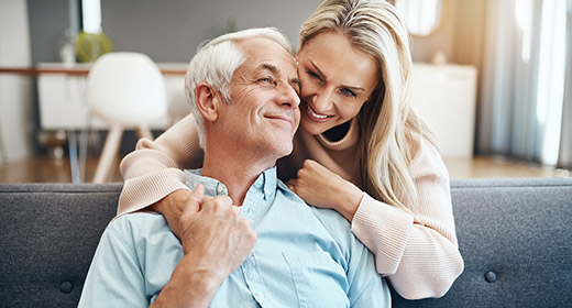 Older man sitting on a couch being hugged from behind by his wife