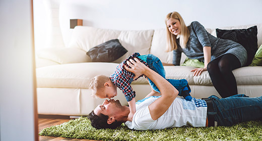 Man laying on a rug playing with his son while the mother watches from the nearby couch
