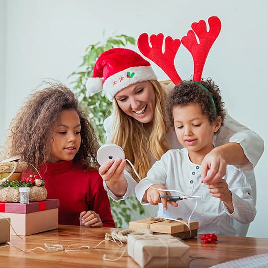 mother wearing a santa hat sitting at a table with her two daughters working on a craft project