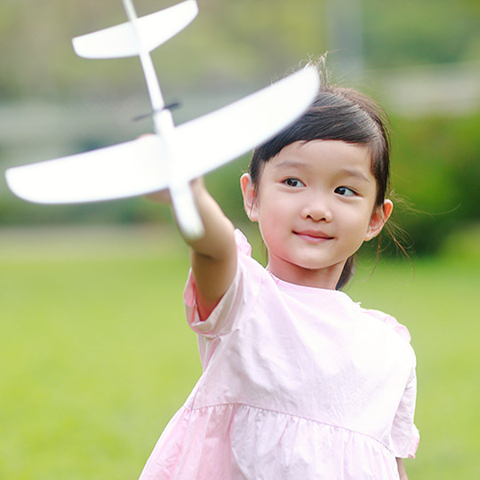 Little girl in a pink dress standing in a grassy park holding out a foam airplane that can be thrown