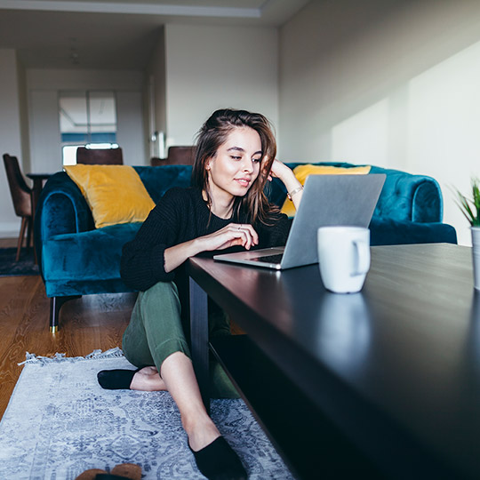 Young woman sitting in front of her couch typing on her laptop that is on a coffee table in front of her