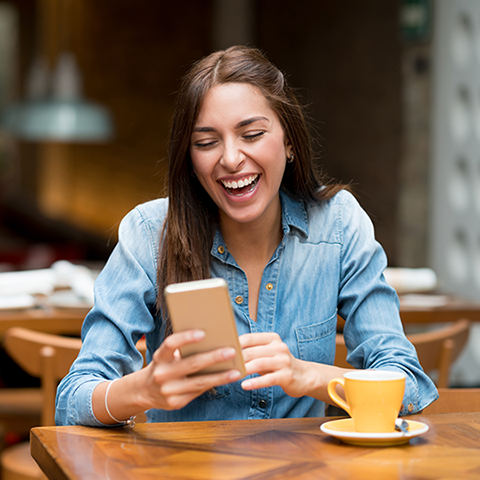/getmedia/950bc999 1d32 4e10 bf2c 76036af62e50/woman at a coffee shop laughing while looking at phone_480x480