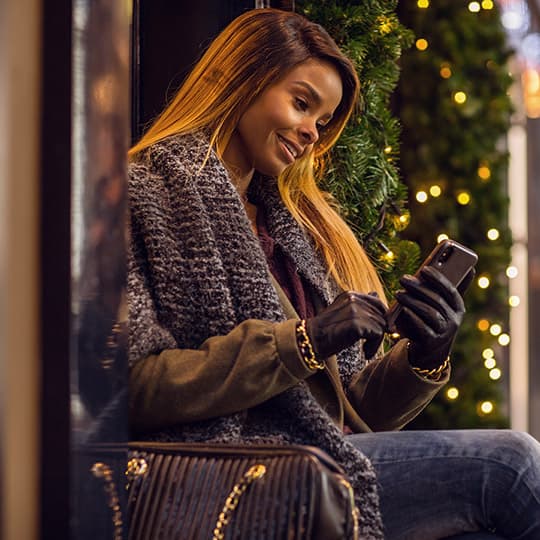 woman in winter coat sitting outside a store and looking at her mobile phone