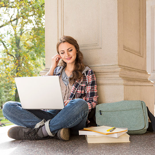College age female sitting against a pillar and looking at her laptop