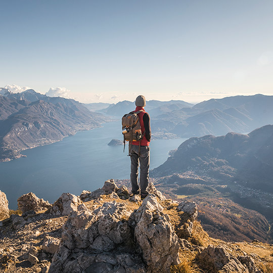 Man in full hiking gear and backpack standing on a cliff peak in a tundra area overlooking a large lake