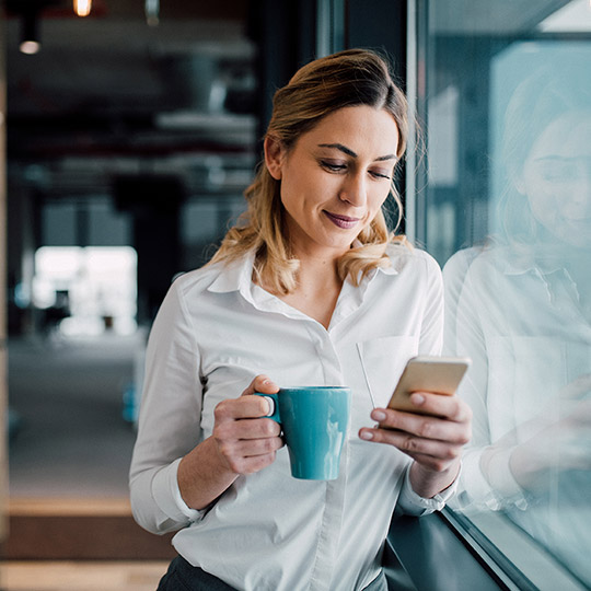 Woman leaning against wall at work looking at her phone while holding a coffee mug