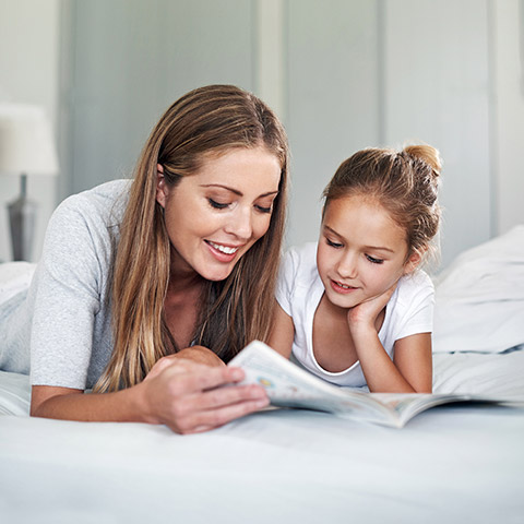 Mother laying on a bed reading to her daughter from a magazine