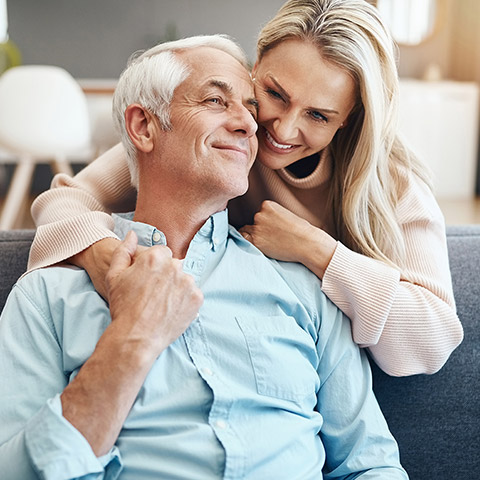 Older man sitting on a couch being hugged from behind by his wife_1