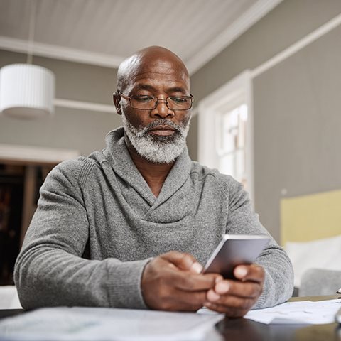 older man wearing glasses sitting at his kitchen table and looking down at his mobile phone