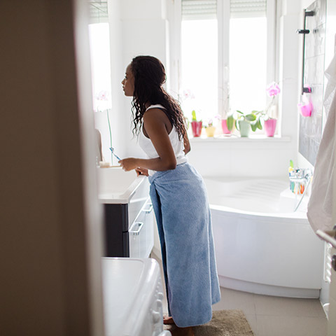 Woman standing at sink in bathroom brushing her teeth after having just taken a shower